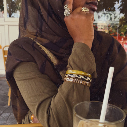 A woman wearing a brown patterned hijab and olive green top sits at a cafe table with an iced coffee in the foreground.