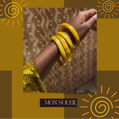 Promotional graphic for 'Summa Bangles Are Here' by Mon Soleil, featuring a close-up of a person's wrist adorned with bright yellow bangles against a patterned brown and beige background, framed by a mustard and brown graphic border with sun illustrations.